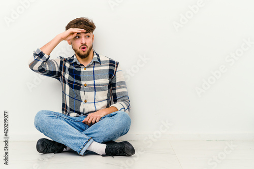 Young Moroccan man sitting on the floor isolated on white background looking far away keeping hand on forehead. © Asier