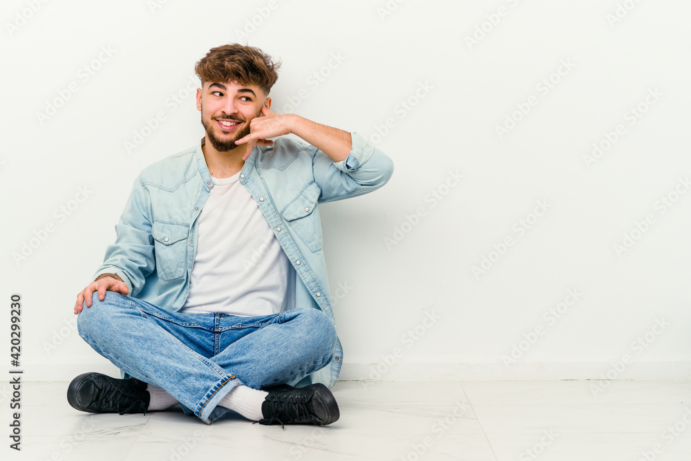 Young Moroccan man sitting on the floor isolated on white background showing a mobile phone call gesture with fingers. Young Moroccan man sitting on the floor isolated on white background showing a mobile phone call gesture with fingers.