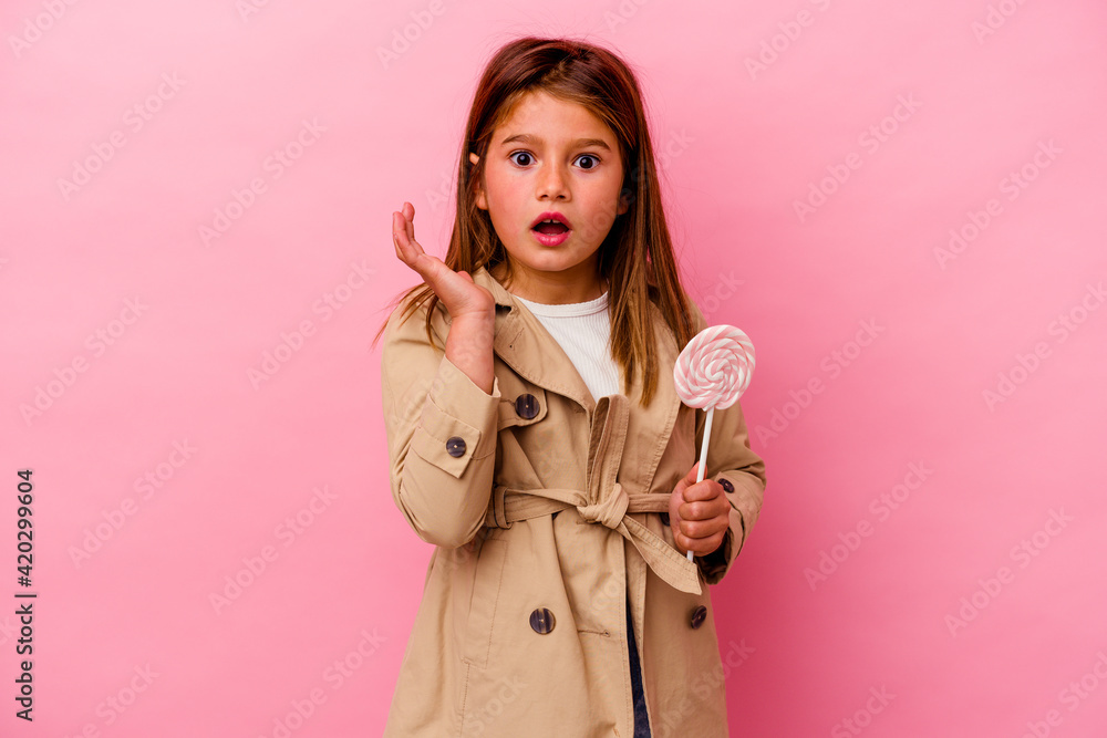 Little caucasian girl holding a lollipop isolated on pink background surprised and shocked. Little caucasian girl holding a lollipop isolated on pink background surprised and shocked.