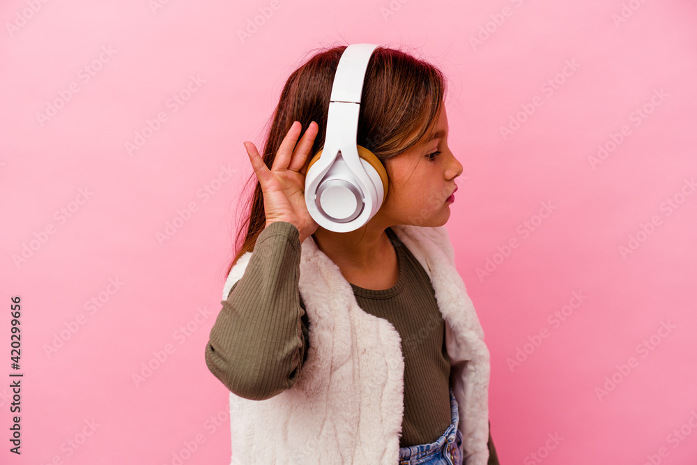 Fototapeta premium Little caucasian girl listening music isolated on pink background trying to listening a gossip.
