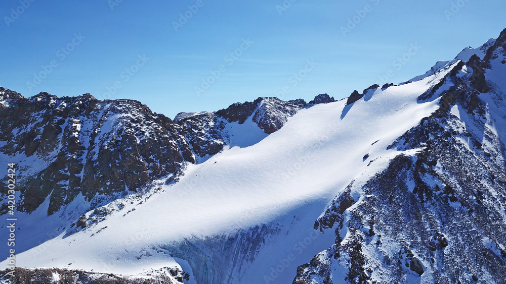 High snow-capped peaks and glaciers. Top view from the drone. Steep cliffs, the wind blows away the snow. There is a lone nunatak. Moraine frozen lake. Climb to the peak. Bogdanovich Glacier, Almaty