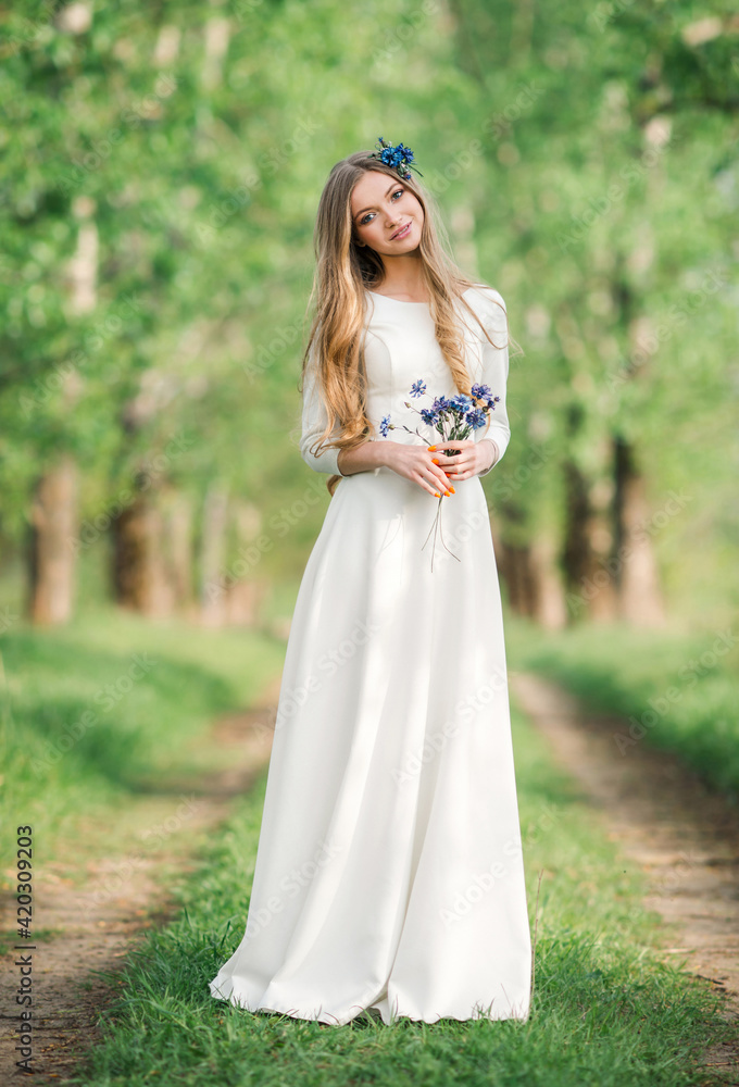 Smiling young woman in white wedding dress with flowers enjoying a summer day in beautiful nature