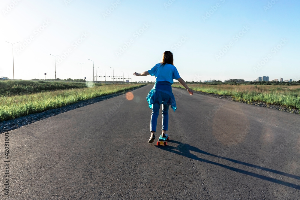 Obraz premium Skater caucasian Young woman from the back in blue jeans and a t-shirt skateboarding on the road in summer, active lifestyle, outdoor activities