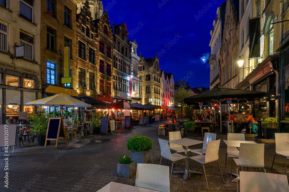 Fototapeta premium Old cozy narrow street with tables of restaurant in historic city center of Antwerpen (Antwerp), Belgium. Night cityscape of Antwerp. Architecture and landmark of Antwerpen