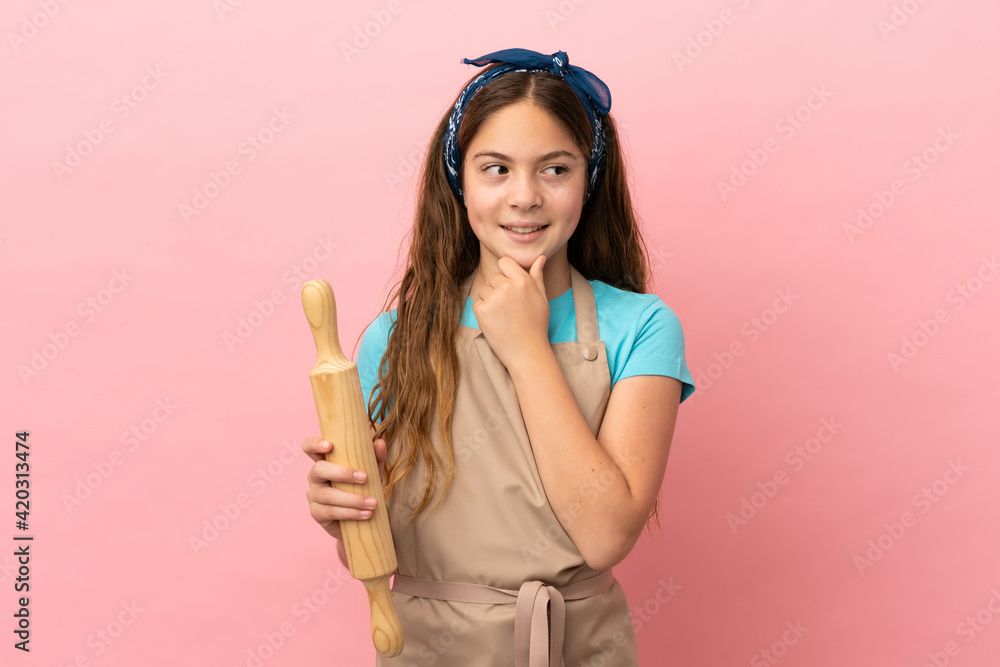 Little caucasian girl holding a rolling pin isolated on pink background looking to the side and smiling Little caucasian girl holding a rolling pin isolated on pink background looking to the side and smiling