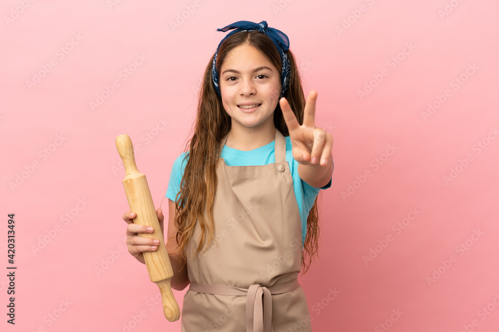 Little caucasian girl holding a rolling pin isolated on pink background smiling and showing victory sign Little caucasian girl holding a rolling pin isolated on pink background smiling and showing victory sign