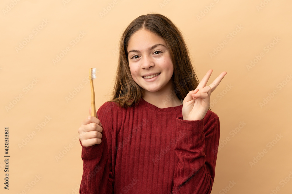 Little caucasian girl brushing her teeth isolated on beige background smiling and showing victory sign Little caucasian girl brushing her teeth isolated on beige background smiling and showing victory sign