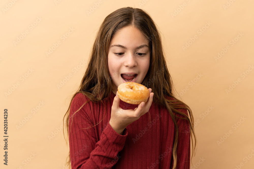 Little caucasian girl isolated on beige background eating a donut