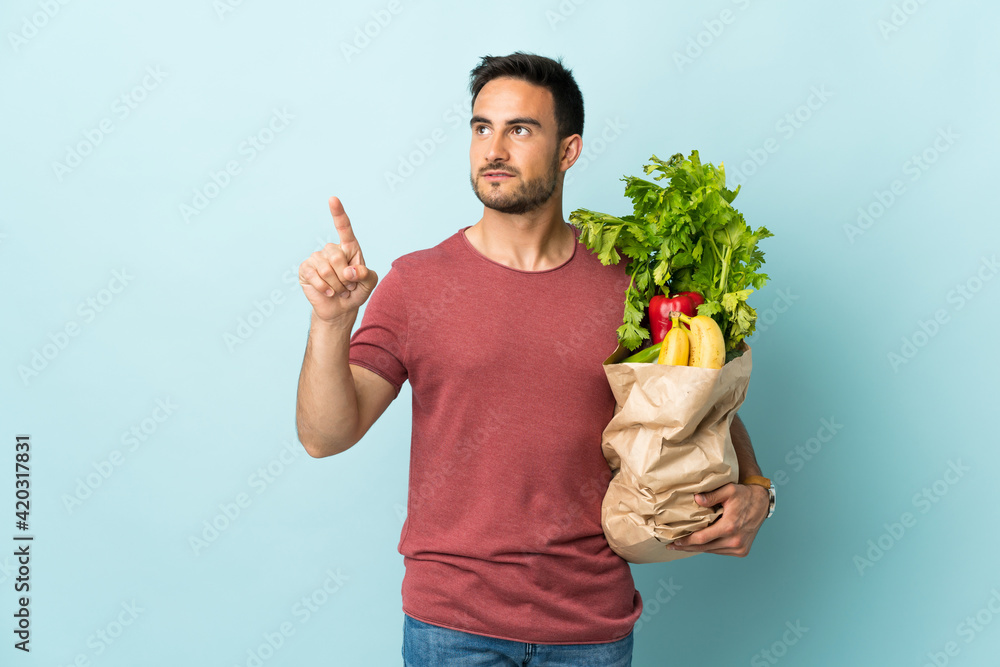 Young caucasian man buying some vegetables isolated on blue background touching on transparent screen Young caucasian man buying some vegetables isolated on blue background touching on transparent screen