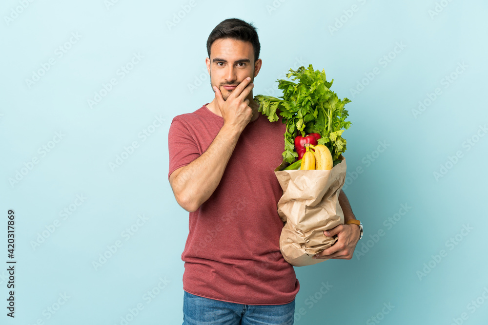 Young caucasian man buying some vegetables isolated on blue background thinking an idea Young caucasian man buying some vegetables isolated on blue background thinking an idea