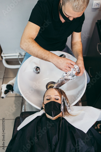 Hairdresser washing hair of a beautiful young adult woman in hair salon. She is wearing protective face mask as protection against virus pandemic.