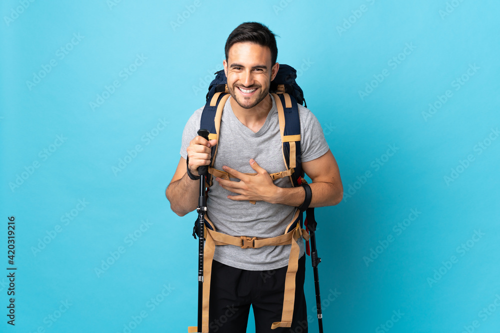 Young caucasian man with backpack and trekking poles isolated on blue background smiling a lot Young caucasian man with backpack and trekking poles isolated on blue background smiling a lot