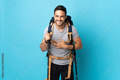 Young caucasian man with backpack and trekking poles isolated on blue background smiling a lot