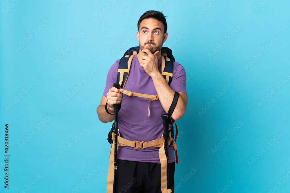 Young caucasian man with backpack and trekking poles isolated on blue background having doubts and thinking Young caucasian man with backpack and trekking poles isolated on blue background having doubts and thinking