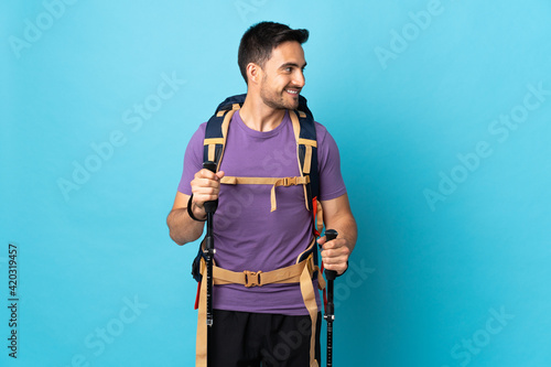 Young caucasian man with backpack and trekking poles isolated on blue background looking side