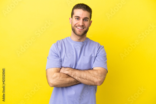 Handsome blonde man over isolated yellow background keeping the arms crossed in frontal position