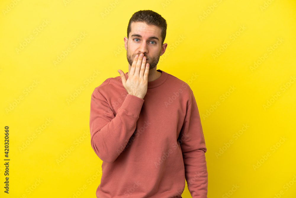 Handsome blonde man over isolated yellow background covering mouth with hand Handsome blonde man over isolated yellow background covering mouth with hand