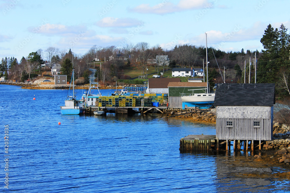 View along the coastline of a Nova Scotia fishing village. Fish sheds, fishing boats, a sailboat on land, lobster traps stacked on a pier.
