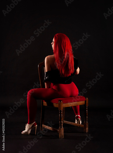 A girl with red hair, wearing red leggings poses on a chair on a dark background.