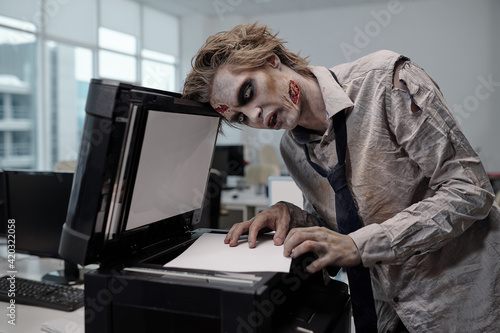 Young spooky businessman with zombie greasepaint on face and hands bending over xerox machine photo