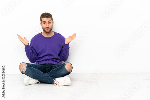 Young man sitting on the floor having doubts while raising hands