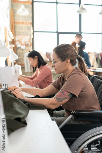 Young disable seamstress sitting in wheelchair by workplace in front of camera and sewing clothes