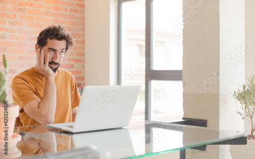 young bearded man feeling bored, frustrated and sleepy after a tiresome, dull and tedious task, holding face with hand and working with a laptop