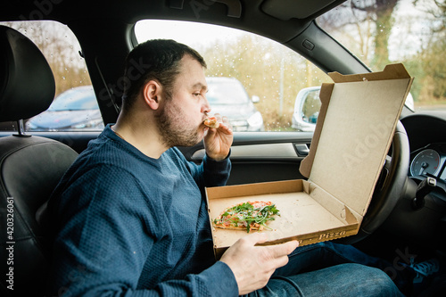 Man with beard eats pizza in his cart. Eat takeaway food in the car. Due to closed restaurants during the pandemic, you are forced to order food. Flatbread in a cardboard box that says "Pizza" 