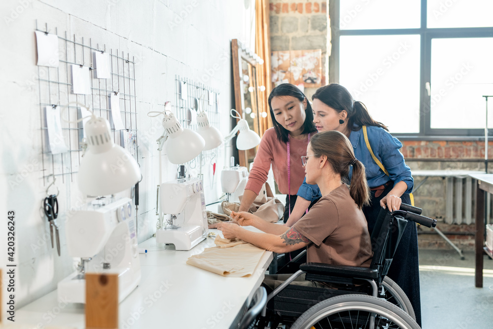 Two young female fashion designers consulting with seamtsress in wheelchair during discussion
