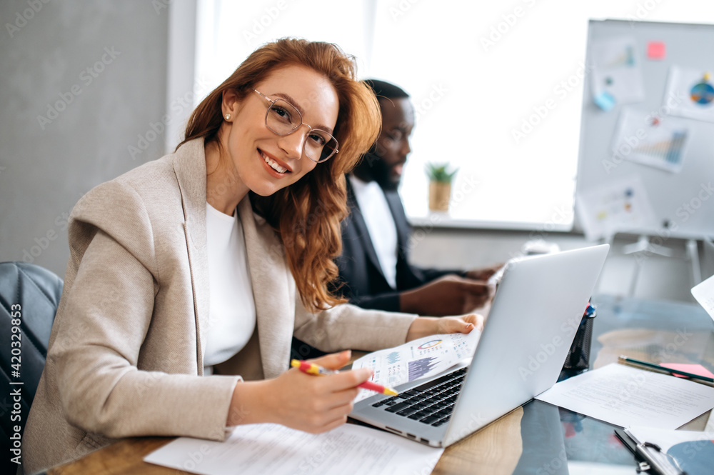 Beautiful redhead woman is using laptop, working on project with colleagues. Happy female employee in stylish suit is sitting in modern office on business meeting, looking at the camera, smiling Beautiful redhead woman is using laptop, working on project with colleagues. Happy female employee in stylish suit is sitting in modern office on business meeting, looking at the camera, smiling