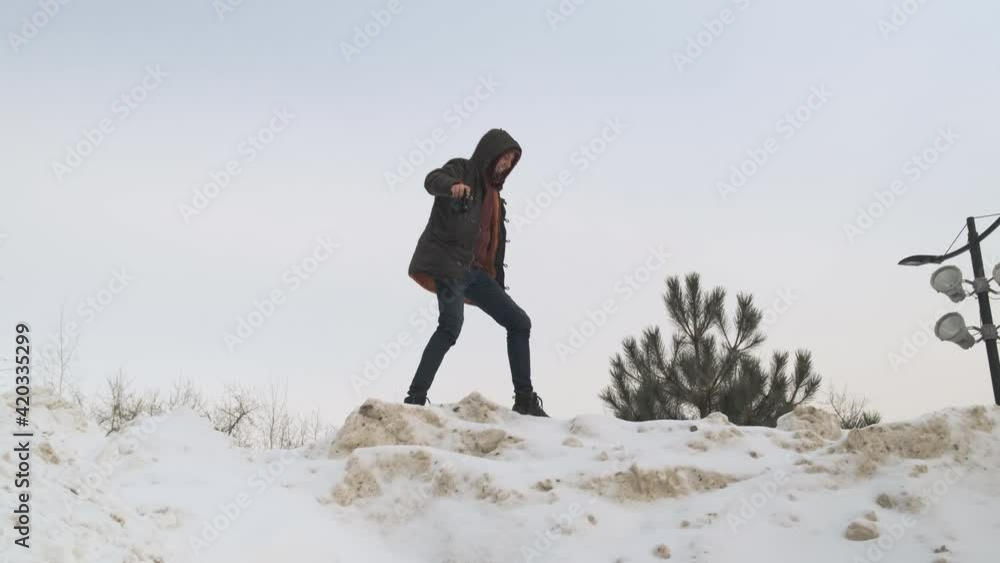 male photographer in black jacket and hood walks along the top of snowy mountain and is ready to take some photos. The naturalist photographer in his hands with camera goes forward and falls comically male photographer in black jacket and hood walks along the top of snowy mountain and is ready to take some photos. The naturalist photographer in his hands with camera goes forward and falls comically