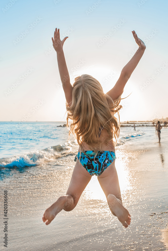 Young happy girl in swimsuit jumps her arms to the sides on the beach against the backdrop of the sea and sunset Young happy girl in swimsuit jumps her arms to the sides on the beach against the backdrop of the sea and sunset