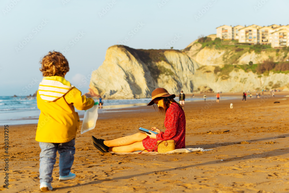 beautiful woman in hat and red dress checking mails from work on the beach in spring easter while her son plays. Holy Week vacations. Spain beautiful woman in hat and red dress checking mails from work on the beach in spring easter while her son plays. Holy Week vacations. Spain