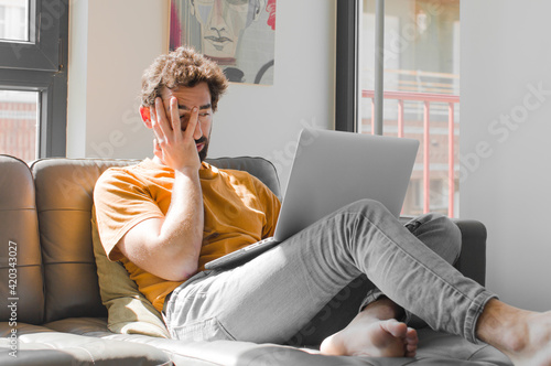 young bearded man feeling bored, frustrated and sleepy after a tiresome, dull and tedious task, holding face with hand with a laptop on a couch laptop concept photo