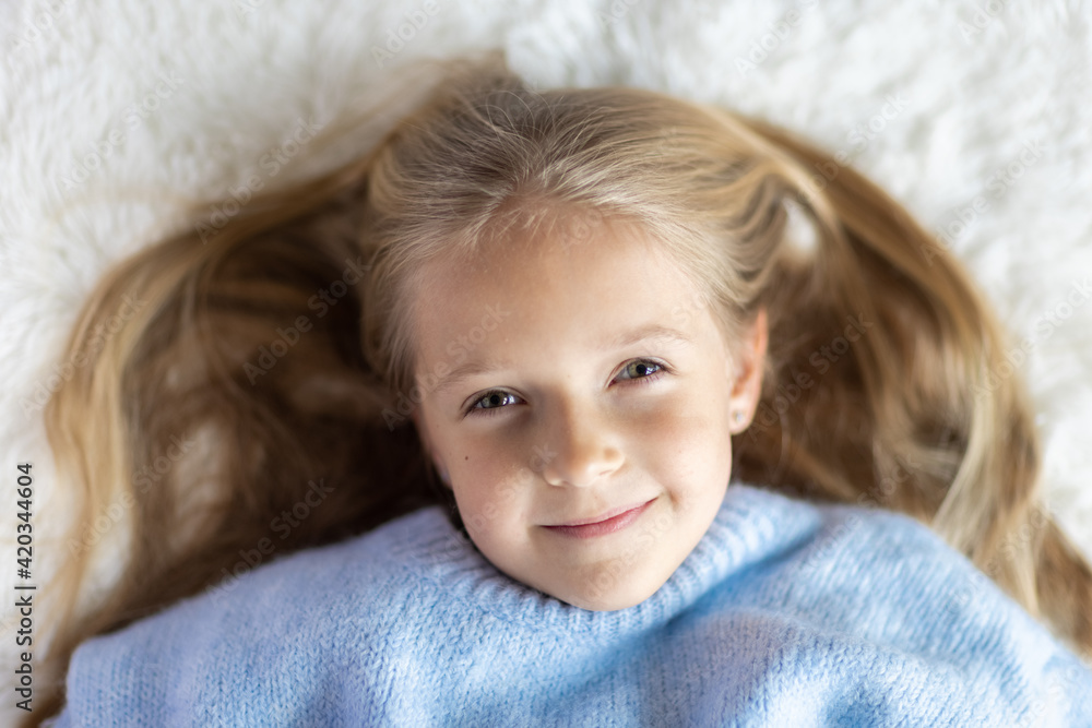 Cute little caucasian girl seven years old with blonde hair lying on bed at home in bedroom. Kid wearing stylish cozy knitted sweater blue color. Happy Child smiling Cute little caucasian girl seven years old with blonde hair lying on bed at home in bedroom. Kid wearing stylish cozy knitted sweater blue color. Happy Child smiling