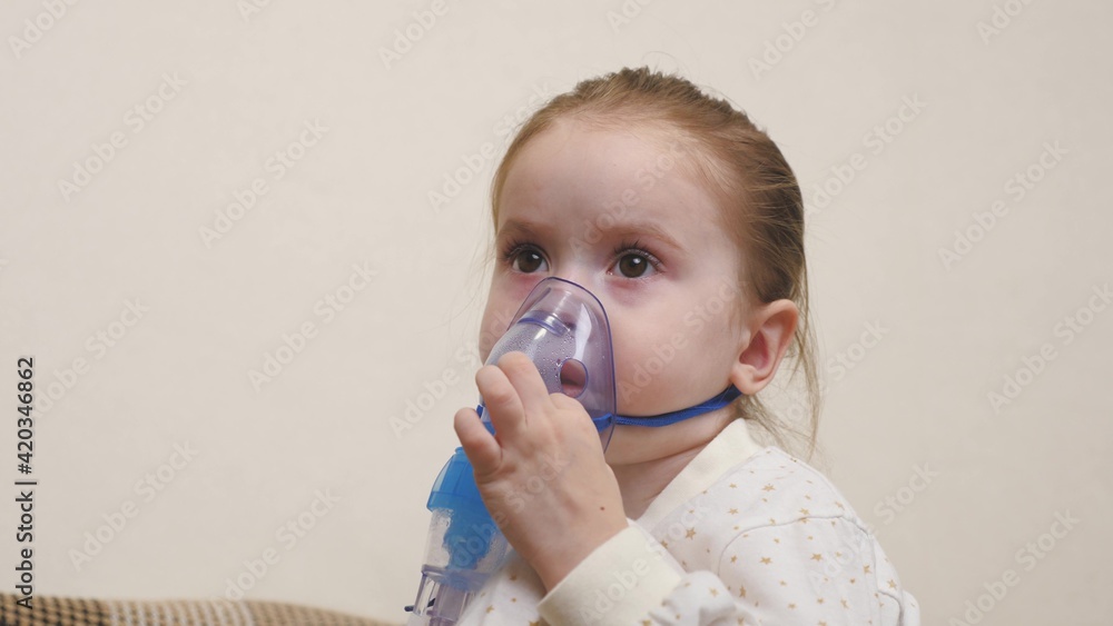 A cute baby in a mask is breathing through an inhaler. Home inhalation procedure. The child is receiving respiratory therapy with a nebulizer. Treat your baby for coronavirus at home. A small child