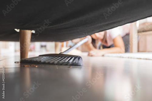 Asian woman cleaning and sweeping dust the floor under the sofa with a broom in the living room. Woman doing chores at home. Housekeeping concept. photo