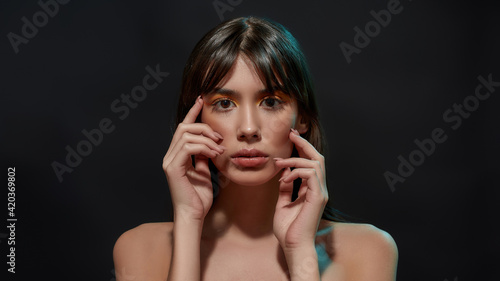 Beauty portrait of young brunette woman with healthy brown hair, big eyes and professional makeup touching her face, posing isolated over black background