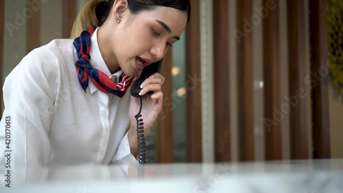 A receptionist woman with uniform at front desk counter in lobby of hotel talking on the phone with guest about the service room. The staff employee working.