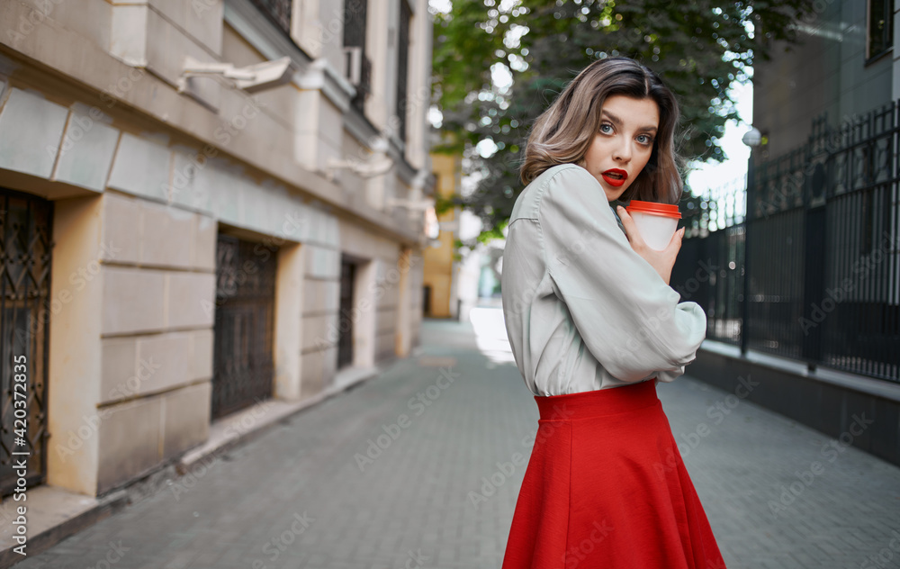 fashionable blonde woman in red skirt stands near the building sidewalk park nature