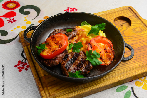 Close up of juicy beef steak served in a pan with bbq grilled tomatoes over traditional Ukrainian tablecloth.