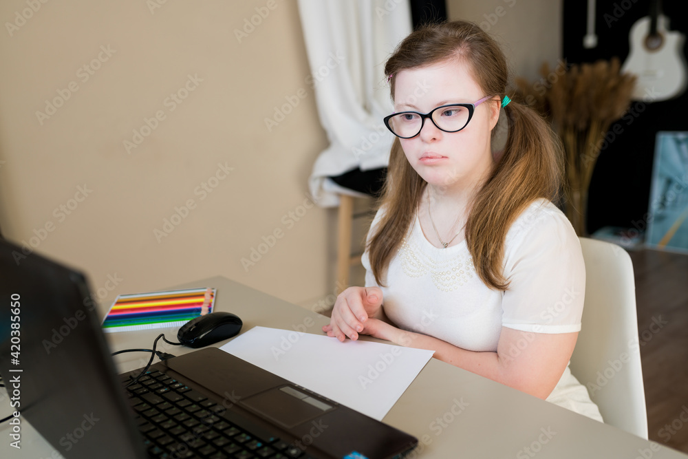 Portrait of teenager girl with Down syndrome learning online at home. Distance learning by video call on laptop. E-learning and online education for kids with special needs concept Portrait of teenager girl with Down syndrome learning online at home. Distance learning by video call on laptop. E-learning and online education for kids with special needs concept