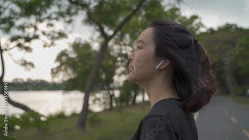 asian sport woman notification from smart watch and answering phone call via wireless earphone at green lake park. female in sportswear talking and hangup phone call by tapping. technology digital.