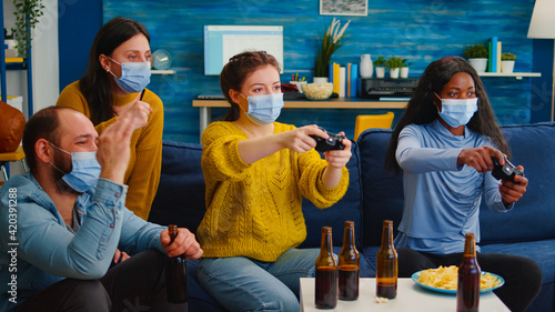 Fototapeta Naklejka Na Ścianę i Meble -  Mixed race women with protection mask playing video game holding wireless controller enjoying new normal party relaxing with friends. Diverse people keeping social distancing against global pandemic