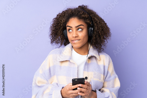 Young African American woman isolated on purple background listening music with a mobile and thinking