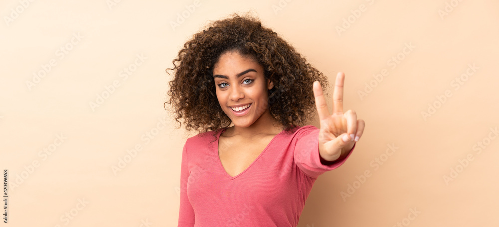 Young African American woman isolated on beige background smiling and showing victory sign