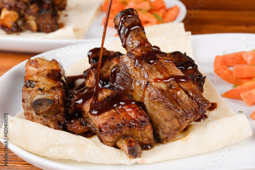 Billede på lærred Sliced grilled pork ribs poured with sauce on pita bread with grilled carrots on a white plate on a wooden background