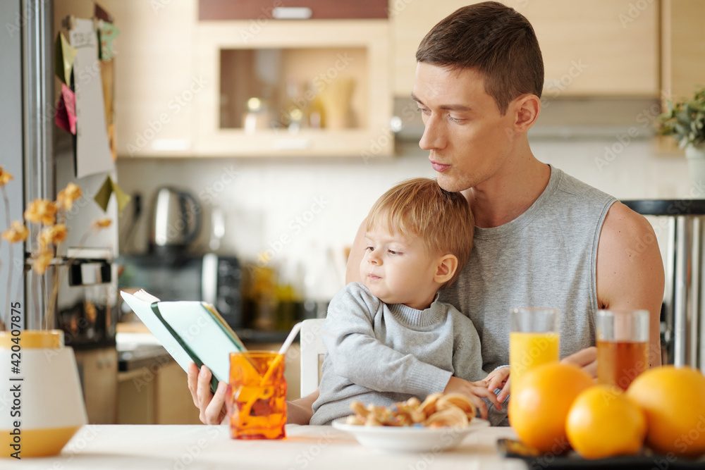 Young father sitting at the table together with his little son and reading him a book during breakfast