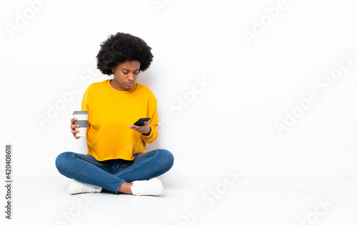 Young African American woman sitting on the floor holding coffee to take away and a mobile © luismolinero