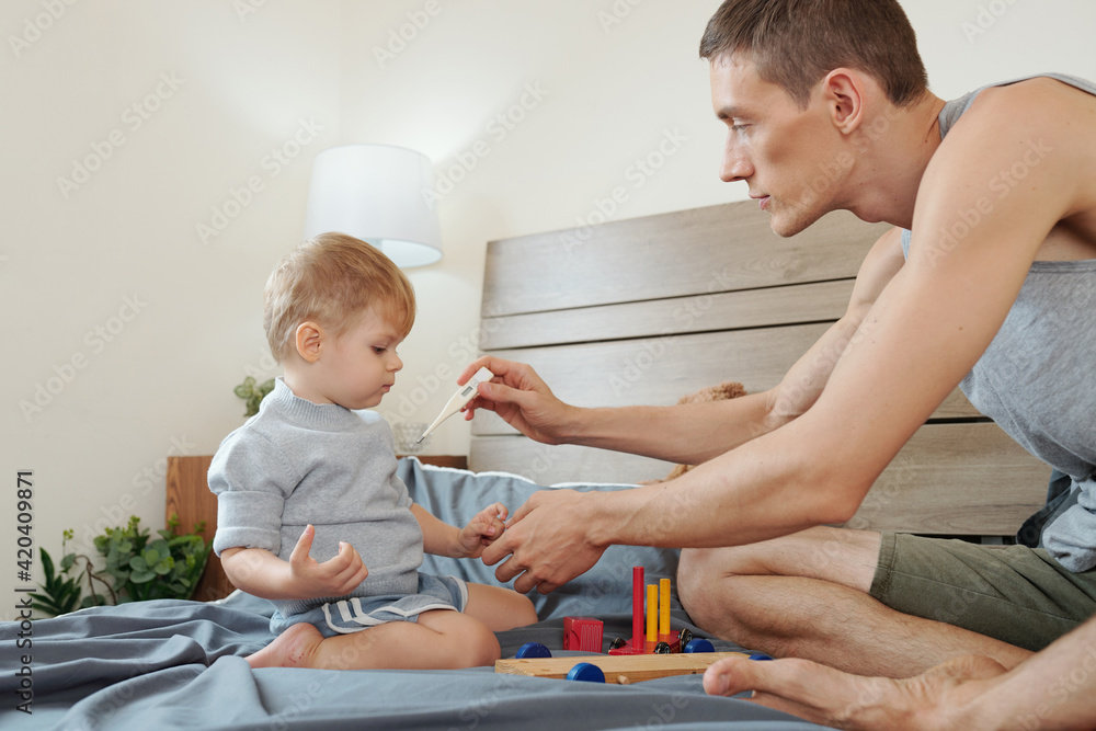 Little boy sitting on bed and playing while his father measuring his temperature during illness Little boy sitting on bed and playing while his father measuring his temperature during illness